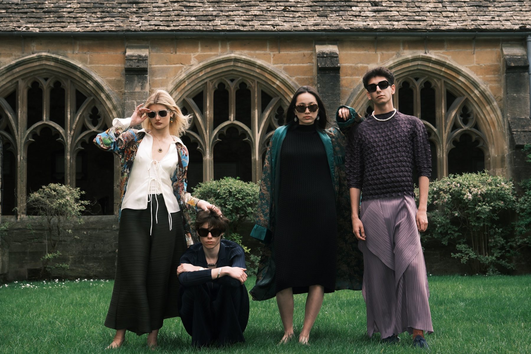 Five models in sunglasses stand and crouch on the lawn of a stone cloister with pointed arched windows, in layered, pleated and patterned clothes.