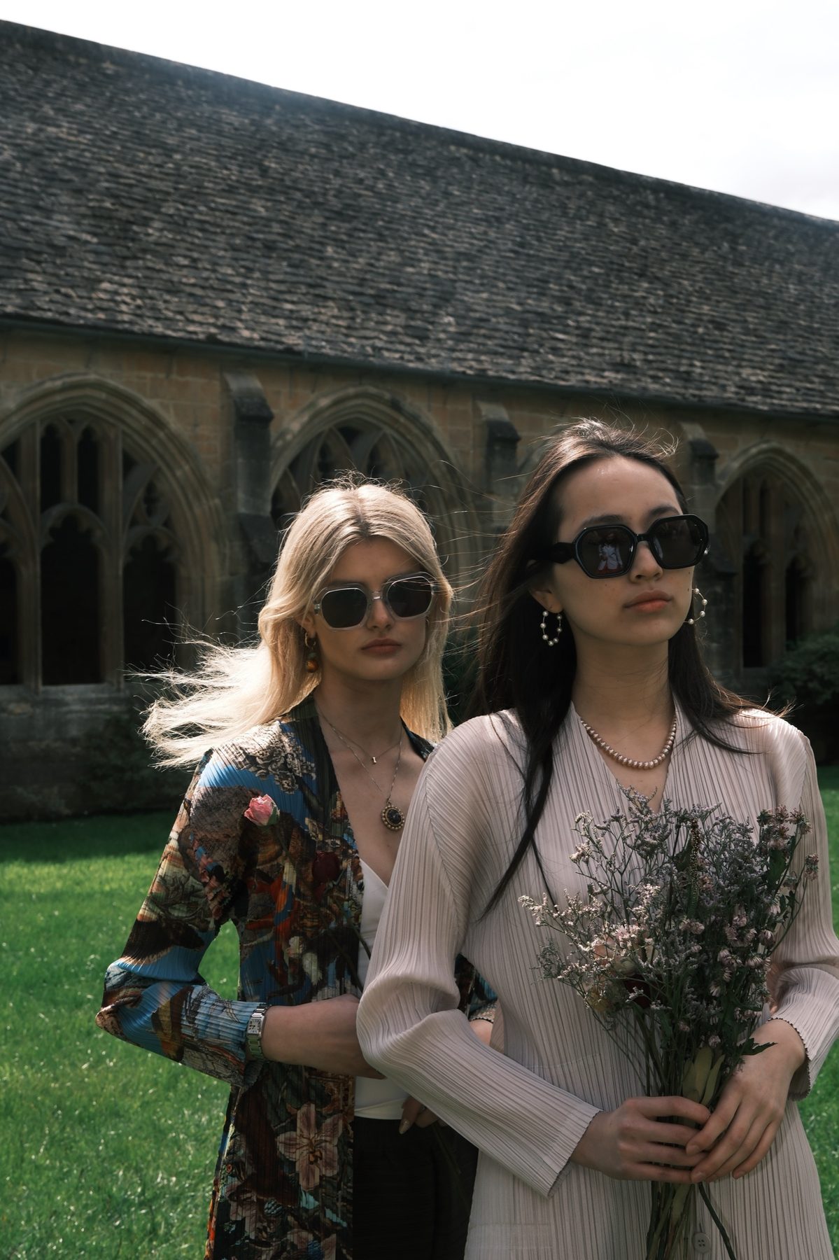 Two models in sunglasses in front of the cloister, one in a patterned jacket, the other in a pale pleated top holding dried flowers.