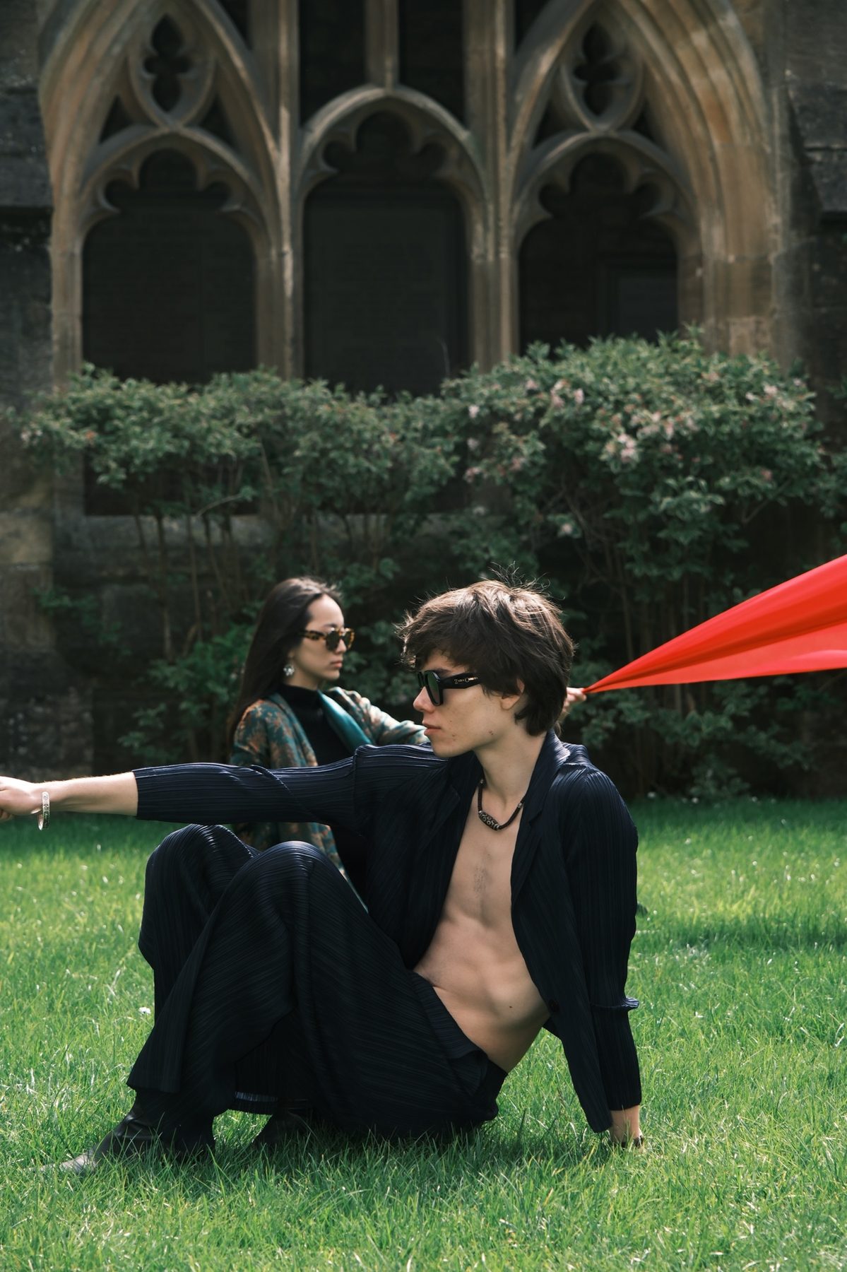 A model in dark pleated clothes sits on the grass, one arm stretched out, holding a red cloth, with another model behind them.