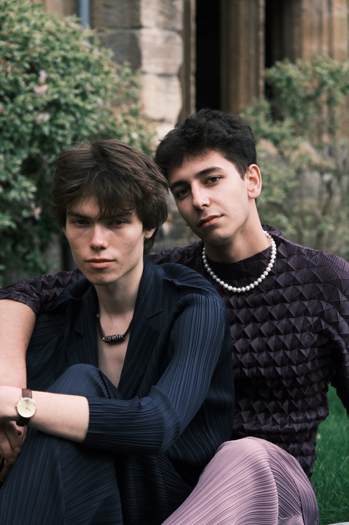 Close portrait of two models seated together on the grass, one in a navy pleated shirt, the other in a purple textured knit and a pearl necklace.