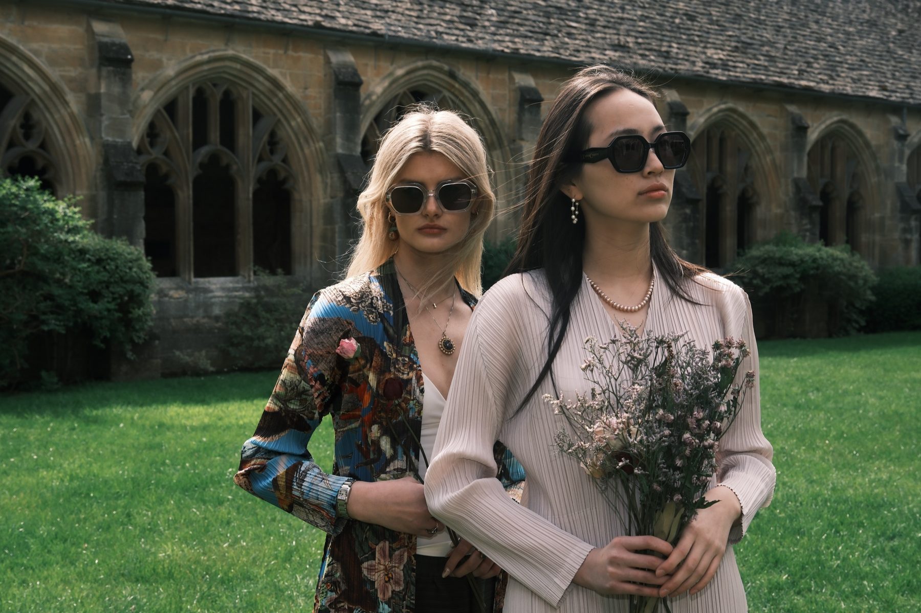 Two models in sunglasses against the cloister's arched windows, in a patterned jacket and a pale pleated top, holding dried flowers.