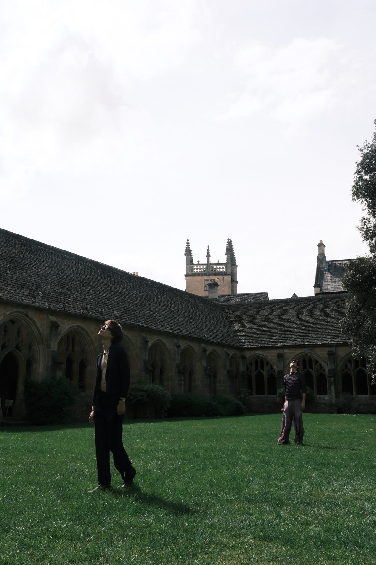 Two models far apart on the lawn, looking up at the sky, with the cloister roof and tower behind them.