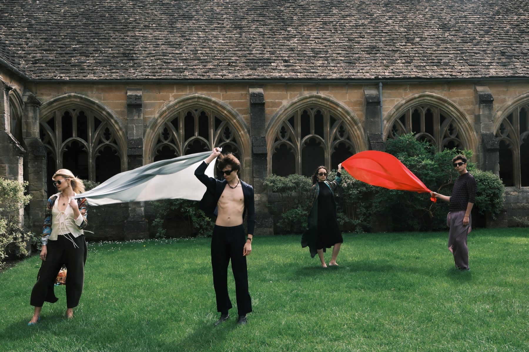 Four models on the lawn holding long pieces of cloth that stream out in the wind, one white and grey, one red.