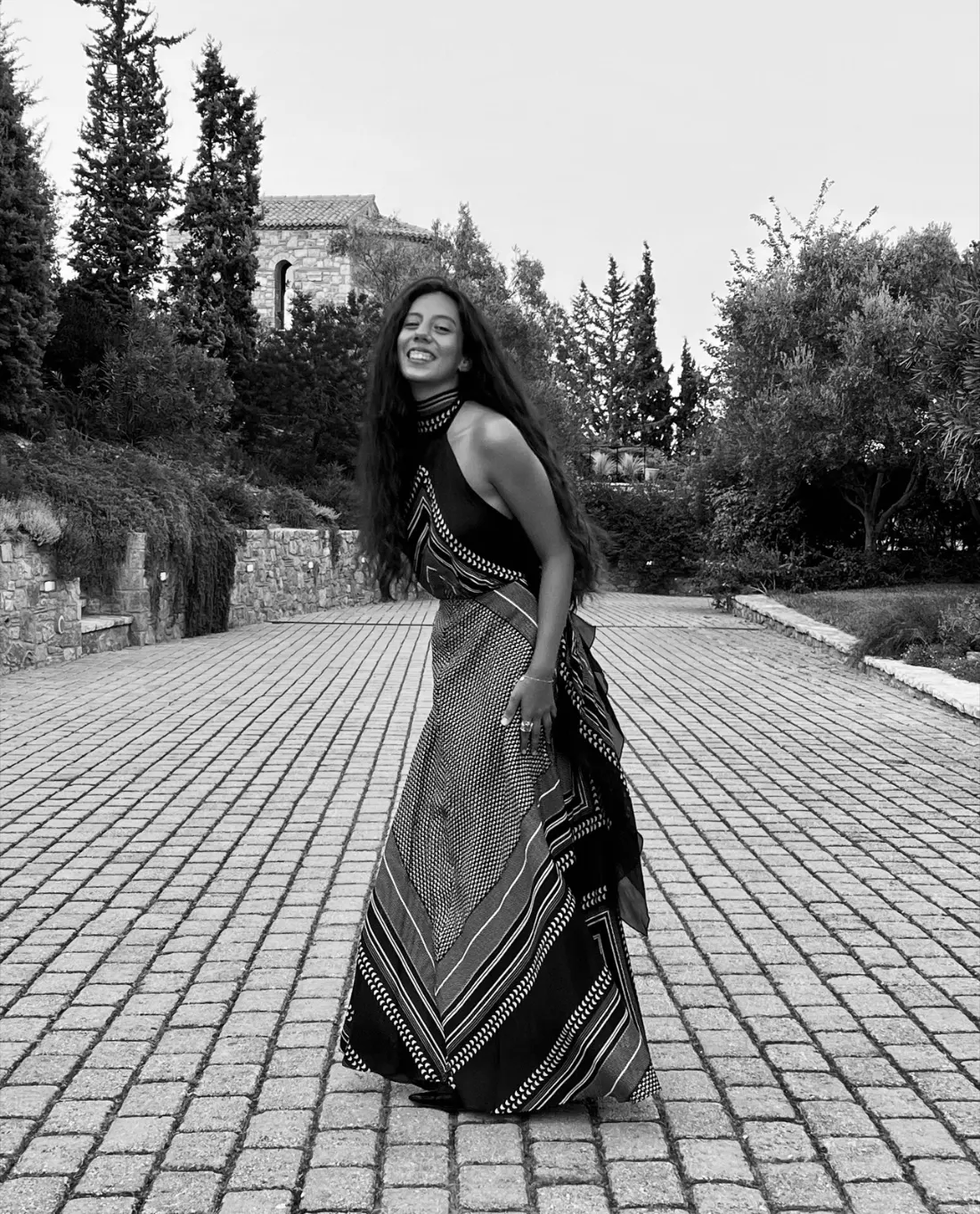 Zaira Christa laughing, mid-turn in a long patterned halter dress on a cobbled path